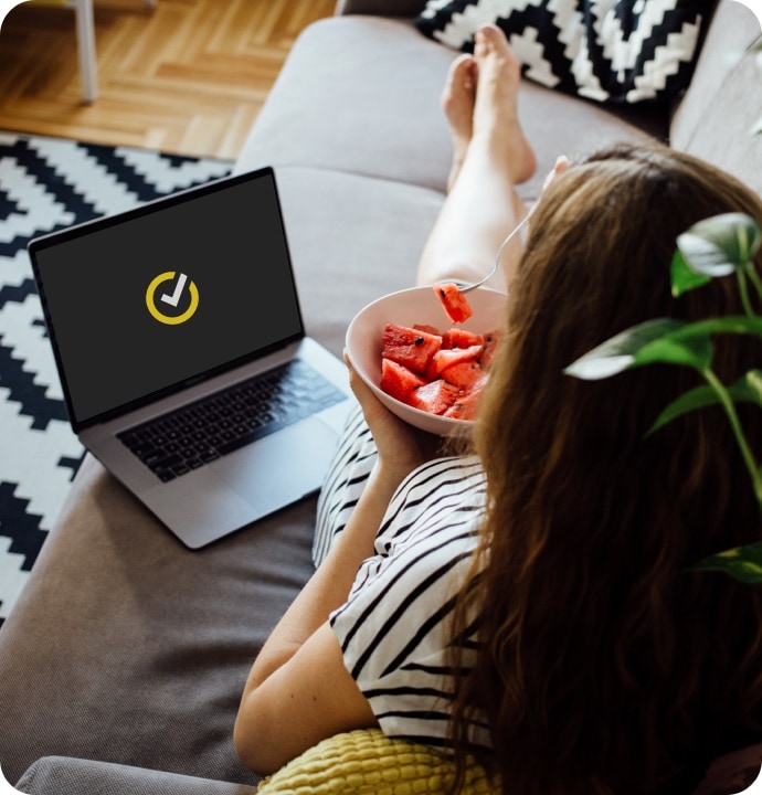 ソファに横たわりフルーツを食べている女性と、ノートンロゴを表示したノートパソコン